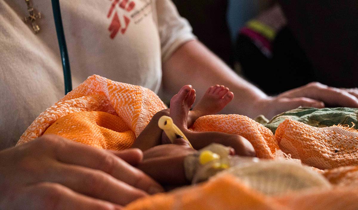 Små spädbarnsfötter sticker upp ut en orange filt med kroppen av en sjukvårdspersonal i bakgrunden