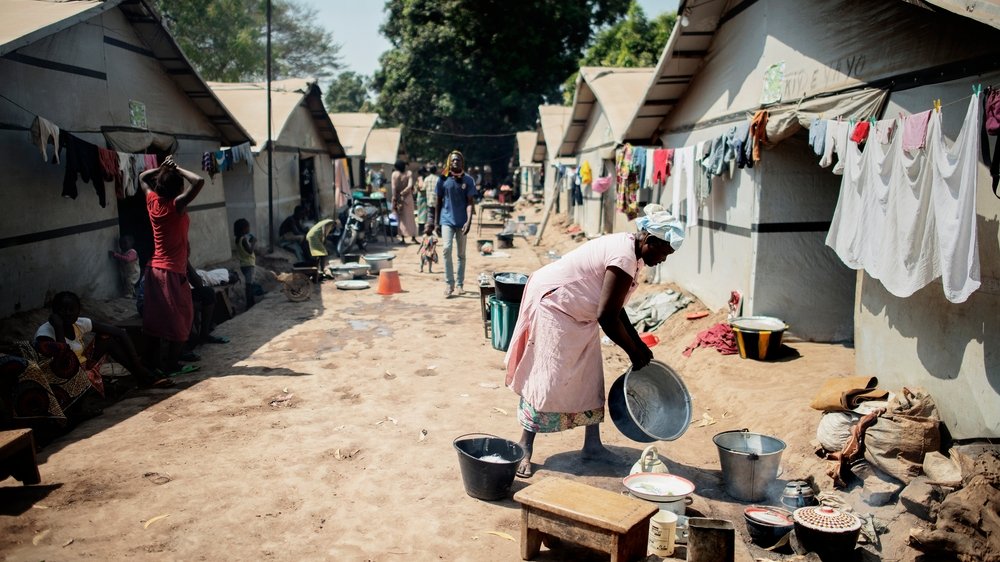 En kvinna diskar stora kärl, i bakgrunden syns ett tältläger och torklinor fulla av tvätt