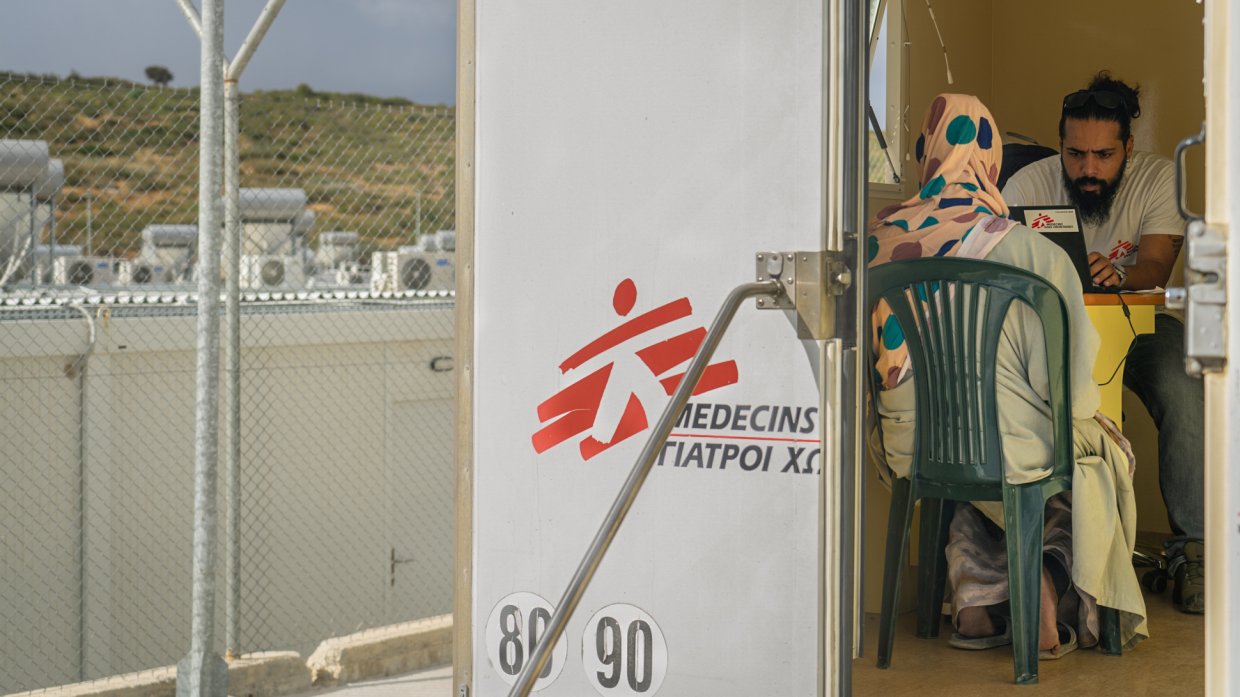 En kvinna sitter på en stol med ryggen mot kameran i en vit container med Läkare Utan Grånskers logotyp på, i en flyktingförläggning på Lesbos