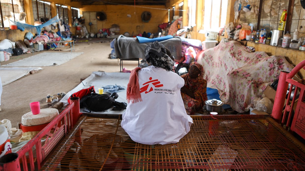 Kvinna med MSF-väst sitter med ryggen mot kameran på en enkel säng i en stökig omgivning.