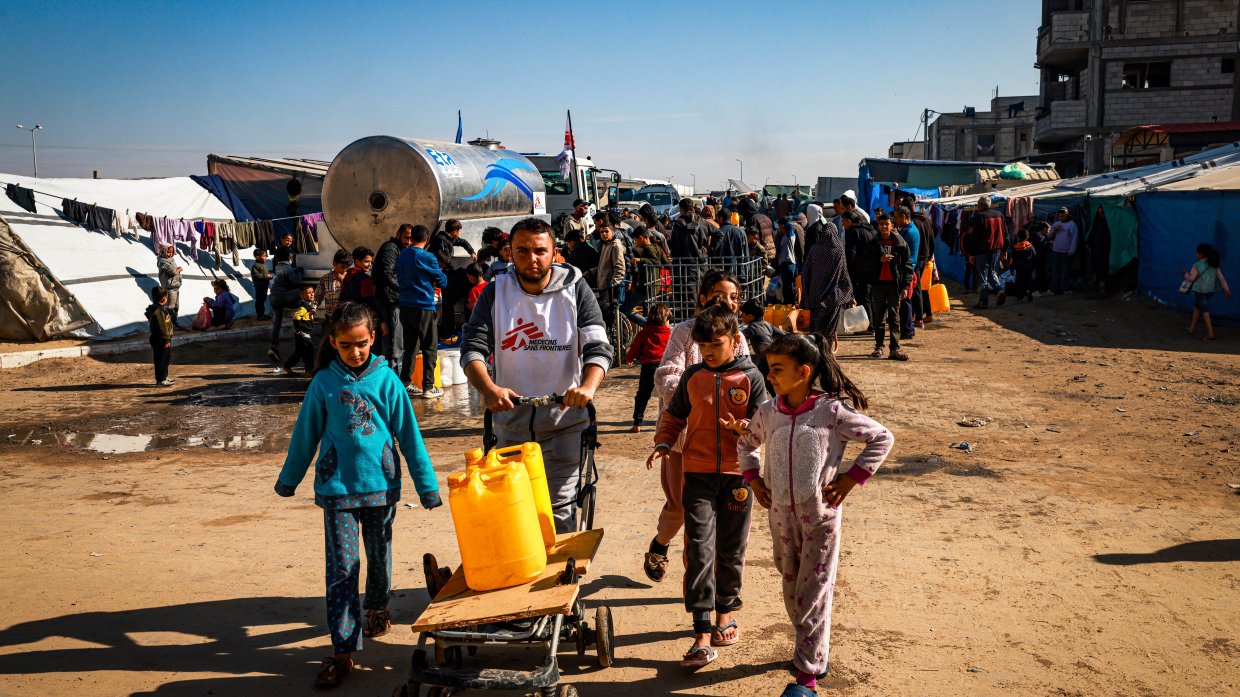 En man i Läkare Utan Gränser väst puttar en vagn med vattendunkar. Bredvid honom går tre barn.