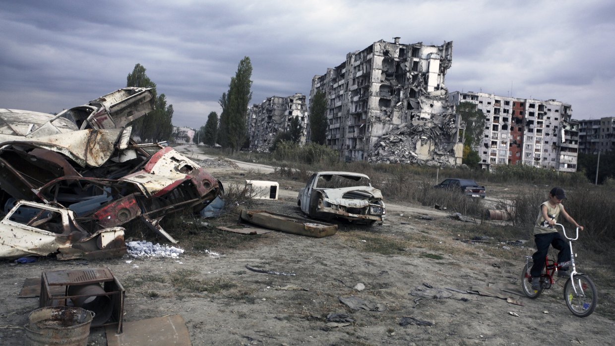 I förgrunden syns en förstörd bil, i bakgrunden ett trasigt hus. 