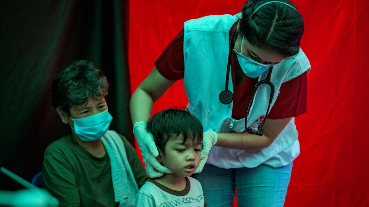 En läkare gör utför en undersökning på ett barn. Barnen sitter i en annan kvinnas knä.