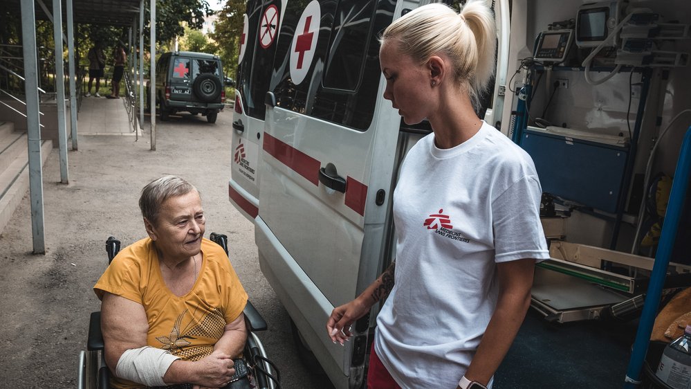 En kvinna står bredvid en ambulans och hjälper en äldre kvinna i rullstol och med bandage runt ena armen.