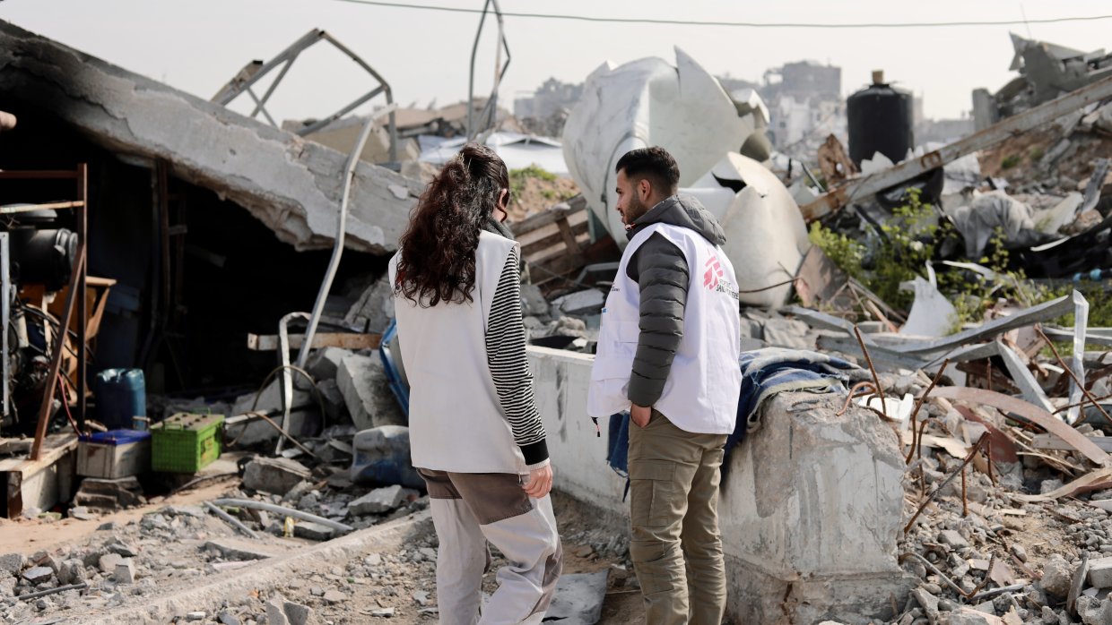 En man och en kvinna från Läkare Utan Gränser står framför rasmassorna av en byggnad som sprängts.