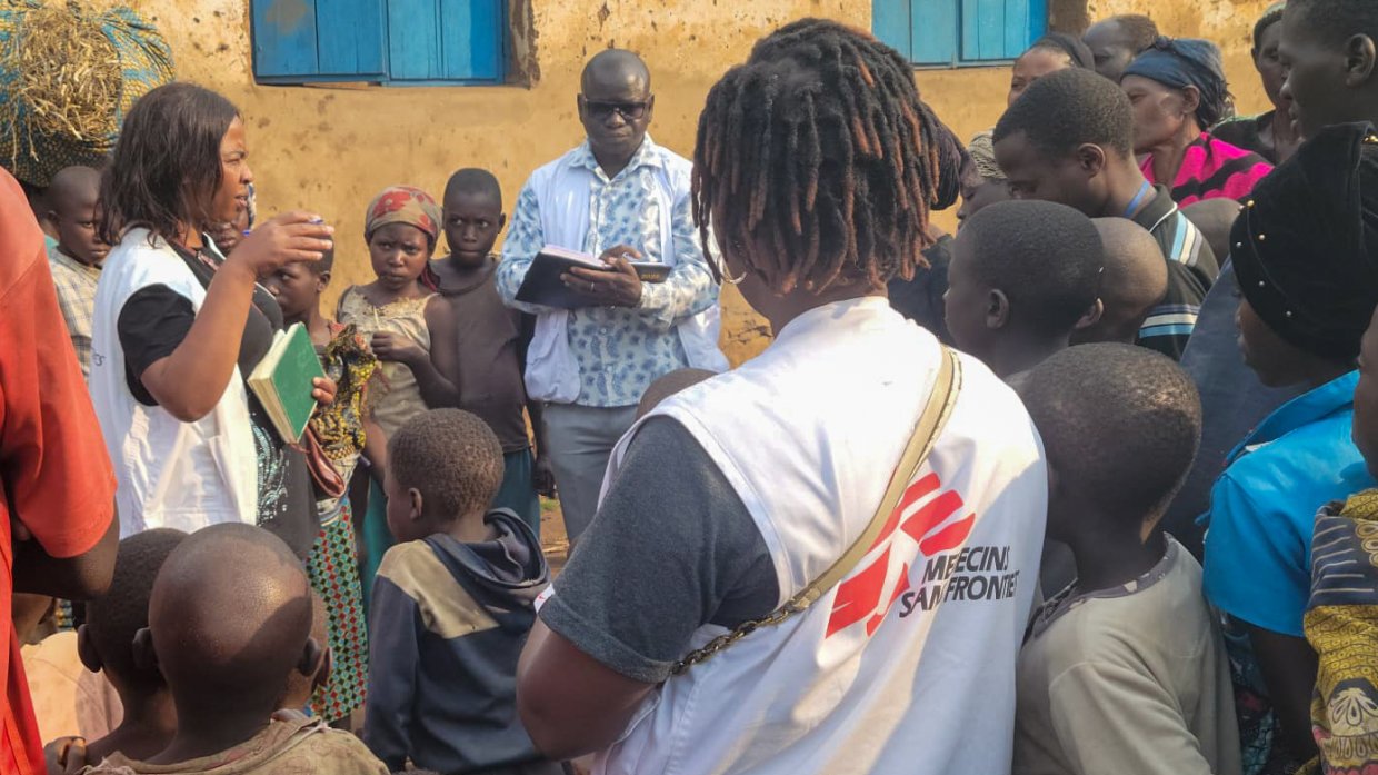 Tre personer i Läkare Utan Gränser-västar står i en cirkel tillsammans med andra civilklädda personer.