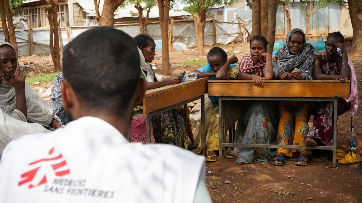 Kvinnor runt ett bord utomhus. En man med Läkare Utan Gränser-väst i förgrunden.