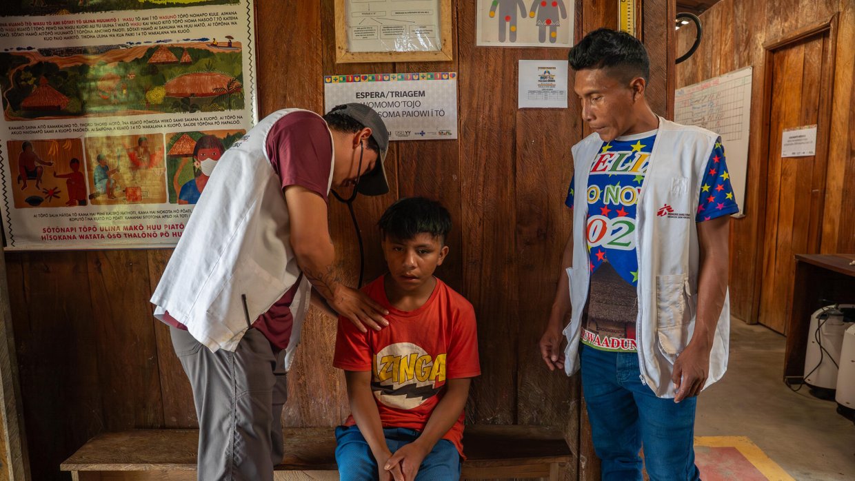Två män i Läkare Utan Gränser-västar och ett barn. Barnet sitter på en bänk, den ena mannen håller ett stetoskop mot barnets rygg och den andra mannen står bredvid.