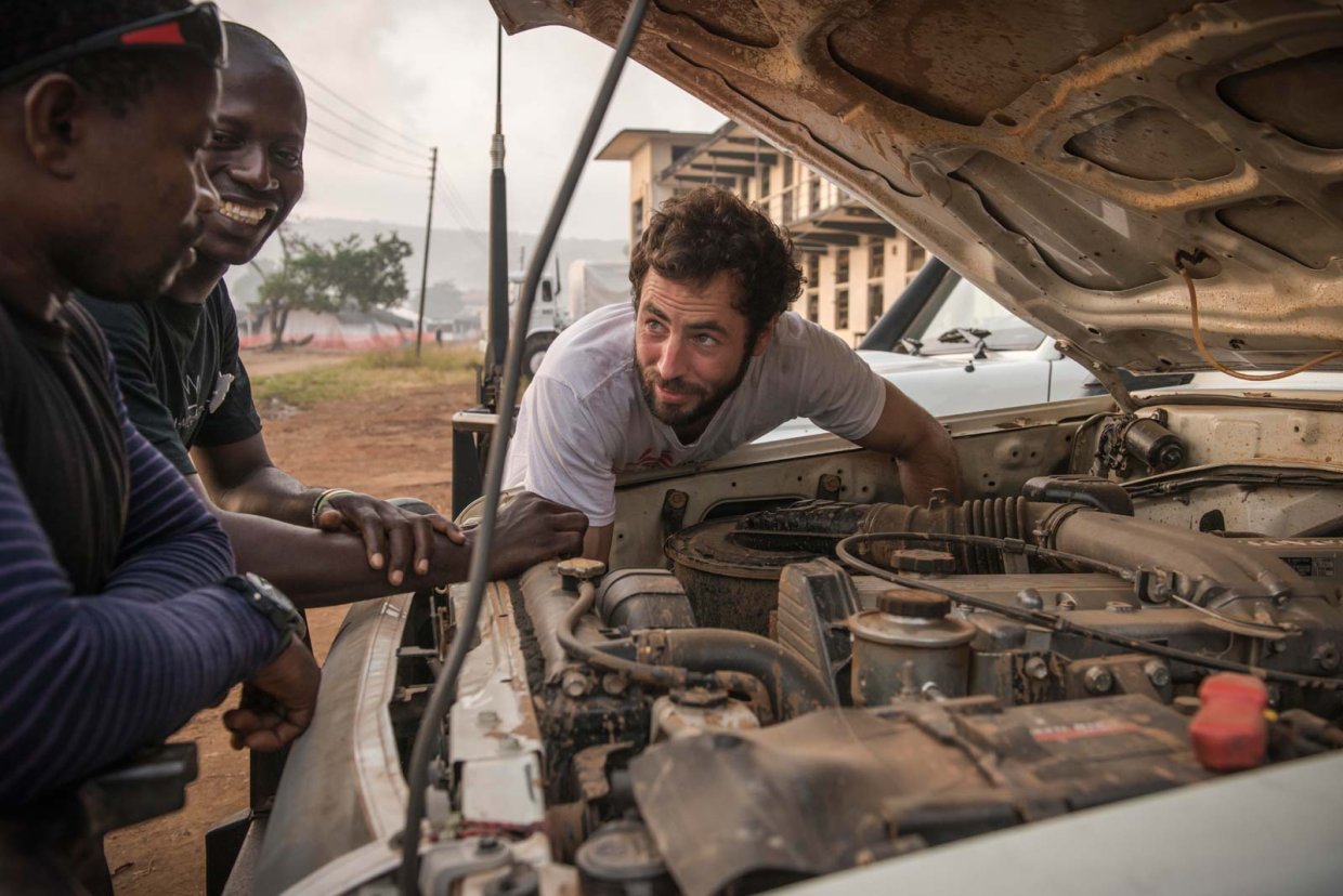 En man mekar med en bilmotor samtidigt som han pratar med två skrattande kollegor.