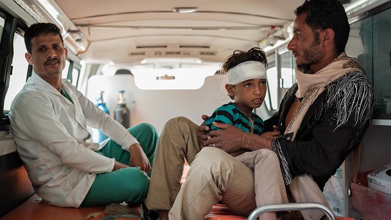 Två vuxna män sitter i bakluckan på en ambulans, den ena mannen har en liten pojke i knät med bandage runt huvudet