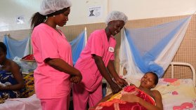 Två sjuksköterskor tittar till en mamma och hennes nyfödda barn som ligger i en sjukhussäng.