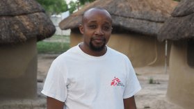 Sjuksköterskan Wilson Thiong'o står i Läkare Utan Gränser-tröja utanför lerhyddor i Sydsudan.