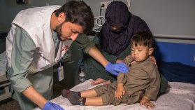 ett barn sitter på en sjukhussäng med sin mamma medan en läkare antecknar i barnets journal 