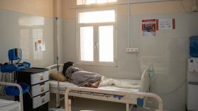 Kvinna ligger på sjukhussäng med ryggen mot kameran, solen lyser in genom fönstret.