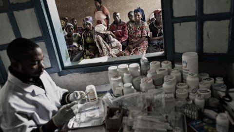 En läkare sitter vid en stor mängd mediciner. Människor väntar utanför ett fönster.