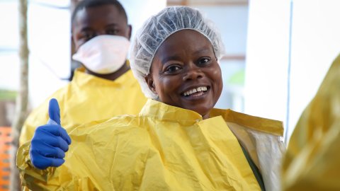 En sjuksköterska i gul skyddsrock och blåa handskar gör tummen upp.