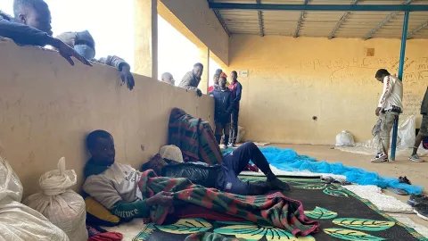 Två män ligger på en filt i ett mottagningscenter i Assamaka, Niger. Runtomkring dem är flera andra män.