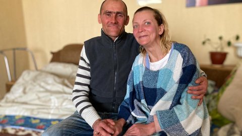 Sergej och Natasha sitter på en säng och håller varandras händer. 