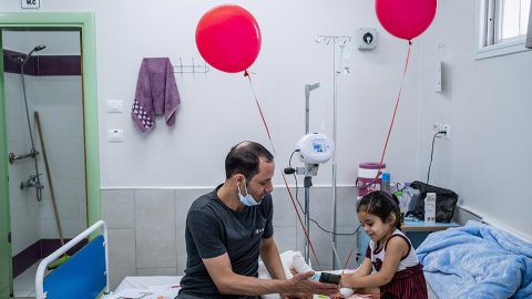Mohammed Aboud sitter brevid sin fyra år gamla dotter Hala som ligger i en säng på barnavdelningen på al-Awda sjukhus i norra Gaza, Palestina. De tittar på en mobiltelefon och två röda ballonger hänger på sängen.