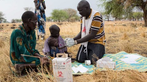 En hälsoinformatör från Läkare Utan Gränser står på knä och testar ett litet barn för malaria utomhus under ett träd i Thaker, Sydsudan. Barnet sitter i en vuxens knä på en filt, bredvid sitter ytterligare en vuxen.