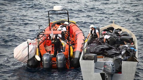 En gummibåt ligger intill en mindre träbåt på centrala Medelhavet. På träbåten ligger flera svarta säckar som två män hjälps åt att lyfta över till gummibåten.