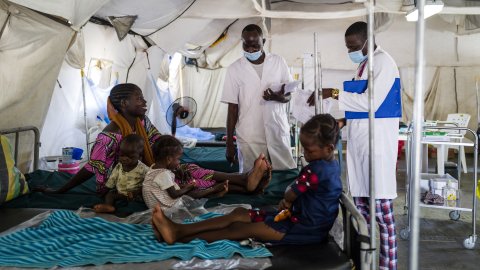 Bria sitter på sängen tillsammans med sina tre barn på sjukhuset i Tukra, Tchad. Bredvid står två läkare.