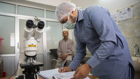 En läkare fyller i en journal. I bakgrunden sitter en patient. 