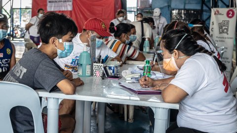 En kvinna i Läkare Utan Gränsers t-shirt sitter vid ett bord. Mittemot sitter en ung kille med munskydd.