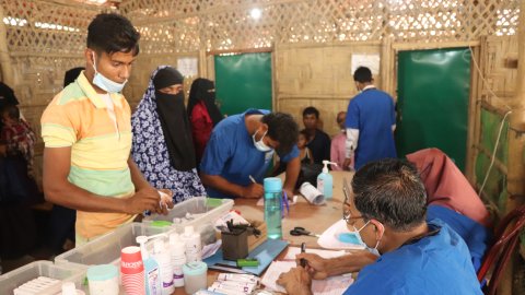 En man står vid ett bord vid vilket en blåklädd man sitter med mediciner framför sig.