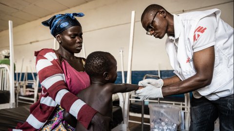 Sjuksköterska undersöker barn som sitter i mammans knä.