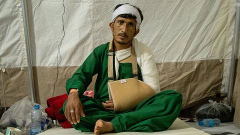 Man med bandage runt armen och huvudet sitter med benen i kors på en sjukvårdssäng och tittar in i kameran.