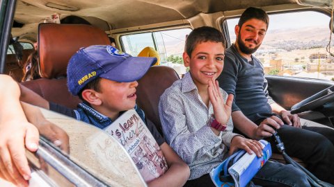 Två pojkar och en man sitter i framsätet av en bil, en pojke vinkar mot kameran och ler