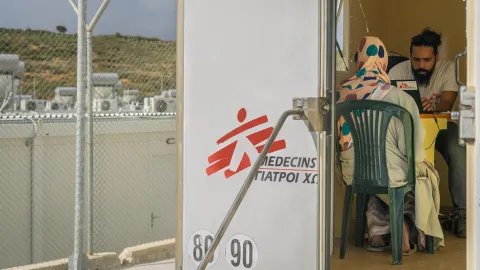 En kvinna sitter på en stol med ryggen mot kameran i en vit container med Läkare Utan Grånskers logotyp på, i en flyktingförläggning på Lesbos