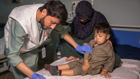 ett barn sitter på en sjukhussäng med sin mamma medan en läkare antecknar i barnets journal 