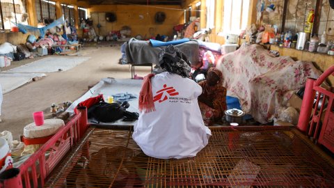 Kvinna med MSF-väst sitter med ryggen mot kameran på en enkel säng i en stökig omgivning.