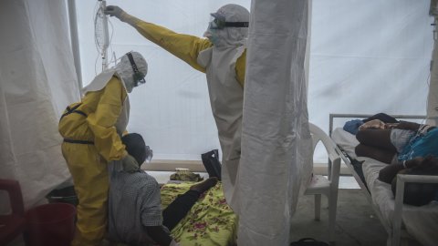 Två personer i heltäckande dräkter vårdar patienter i ett tält.