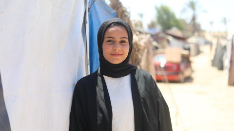 en ung kvinna med svart sjal står vid ett tält och tittar in i kameran