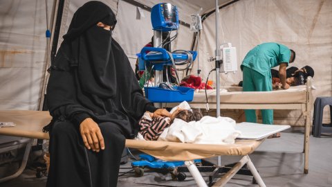 en svartklädd kvinna sitter på en sjukhussäng bredvid sitt sjuka barn