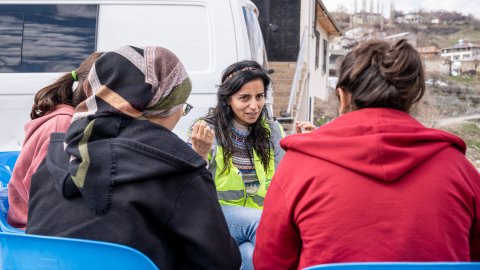 En grupp kvinnor, varav en har gul väst, sitter utomhus och pratar.