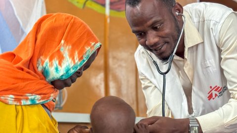 En man med stetoskop och Läkare Utan Gränser-väst undersöker ett barn. En kvinna är med barnet.