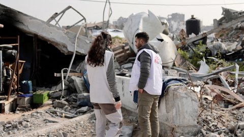 En man och en kvinna från Läkare Utan Gränser står framför rasmassorna av en byggnad som sprängts.