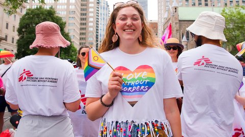 Kvinna med prideflagga och ett hjärta på sin t-shirt med texten Love without borders flankerad av två personer med ryggen till och Läkare utan gränser-västar
