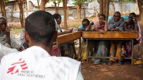 Kvinnor runt ett bord utomhus. En man med Läkare Utan Gränser-väst i förgrunden.