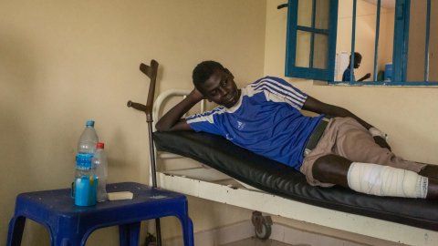 En man i blå tröja på en sjukhussäng. Bredvid sängen står en krycka lutad mot väggen.