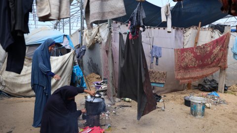 Två kvinnor som lagar mat utomhus. Kläder och tyger hänger på tork ovanför dem.