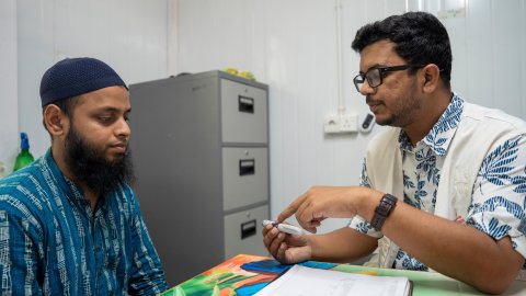 En man med vit Läkare Utan Gränser-väst vid ett bord med en man i blå kläder. Mannen i väst håller upp något som han visar för mannen i blått.
