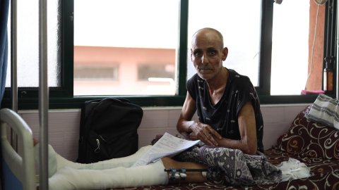 En man med bandagerade ben sitter i en säng och tittar mot kameran.