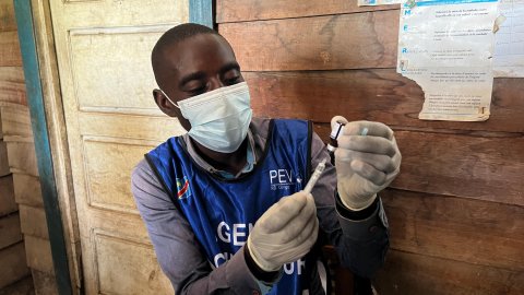 En man med munskydd och handskar håller i en spruta. 