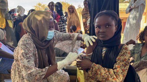 Ett barn gör en grimas medan hon får en spruta av en kvinna med munskydd och handkar som sitter bredvid på marken.
