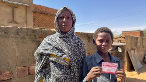 En kvinna och ett barn står och kollar in i kameran. Barnet håller i ett kort med text på.